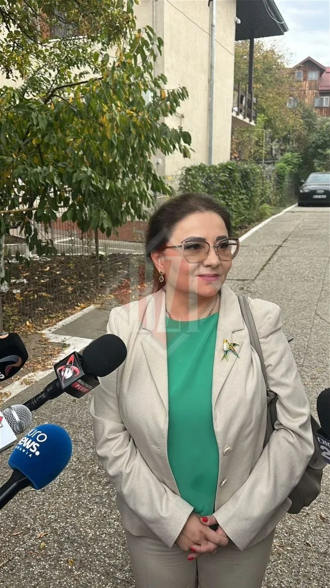 O femeie cu ochelari, purtând un sacou deschis peste un tricou verde, stă în fața mai multor microfoane de presă pe un trotuar.