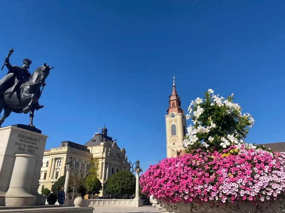 O statuie a unui călăreț pe un piedestal stă lângă un clădire mare și un turn sub un cer senin, lângă un ghiveci cu flori roz și albe.