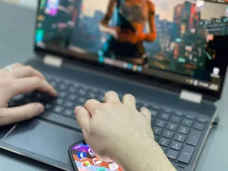 Mâinile unei persoane tastând pe un laptop deschis și ținând un smartphone cu diverse aplicații afișate pe ecran.