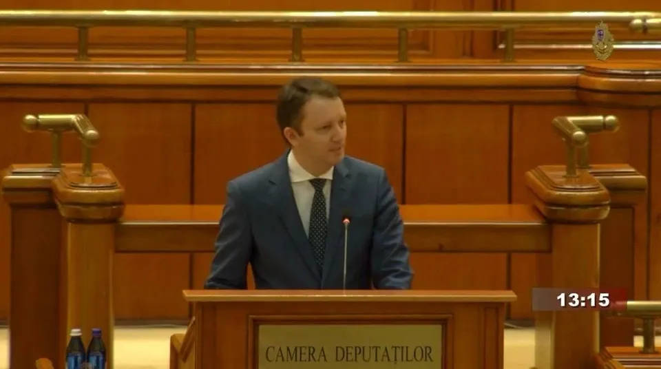 Un bărbat într-un costum albastru și cravată stă în spatele unui pupitru de lemn în fața unui panou cu inscripția Camera Deputaților.