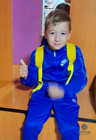 Un copil cu părul scurt și haine sport albastre poartă un rucsac galben și face semn cu degetul mare în sus.