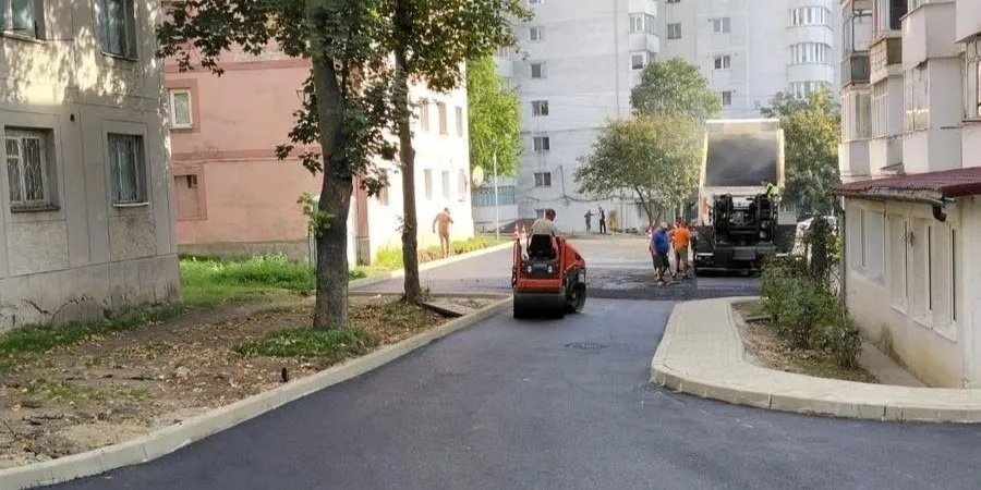 O mașină de asfaltare roșie și un camion negru lucrează pe un drum proaspăt asfaltat lângă clădiri de apartamente și copaci.