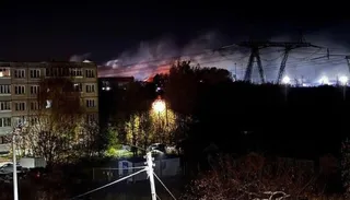 O clădire cu ferestre iluminate stă lângă copaci sub un cer nocturn unde se ridică fum deasupra unor structuri metalice și linii electrice.
