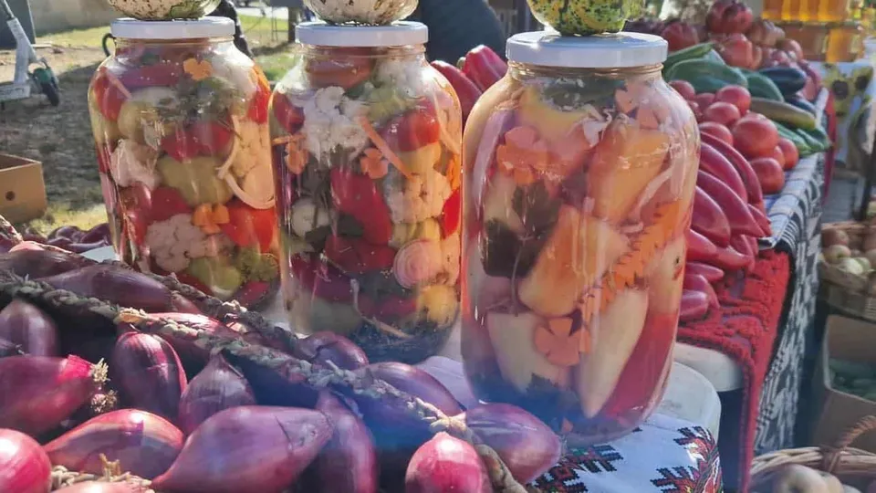 Trei borcane mari cu legume conservate sunt așezate pe un masa lângă diverse legume proaspete, precum ceapă roșie și ardei.