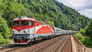 Un tren cu o locomotivă roșie și gri tractează vagoane albastre pe o cale ferată lângă un versant împădurit.