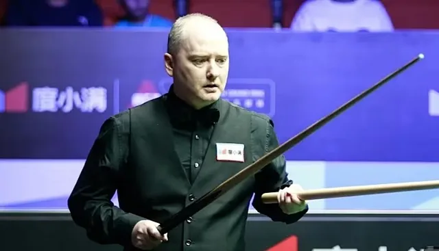 Un bărbat cu vestă neagră și cămașă neagră ține două bâte de snooker în poziție în fața unui fundal albastru.