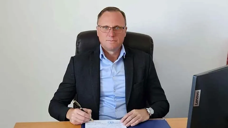 Un bărbat cu ochelari și sacou negru stă la un birou lângă un monitor, ținând un pix deasupra unui document.