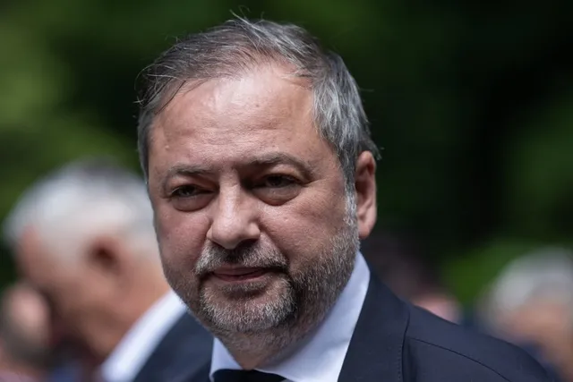 Un bărbat cu părul cărunt și barbă scurtă poartă un sacou închis la culoare și o cămașă albă sub un fundal verde neclar.