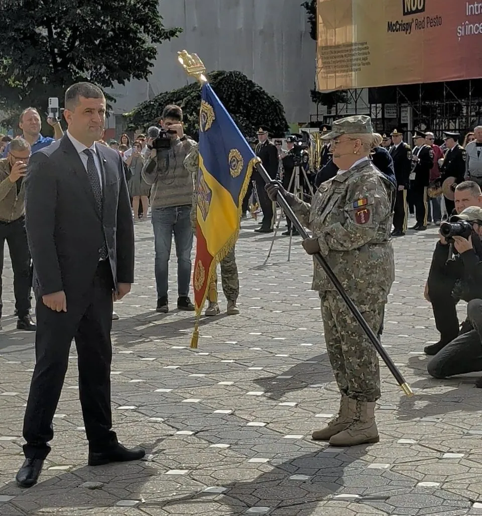 Un soldat în uniformă de camuflaj ține un steag colorat cu un stârf lung în fața unui bărbat într-un costum gri.