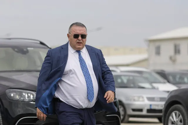 Un bărbat corpolent, cu ochelari de soare, costum și cravată, merge într-o parcare plină de mașini, cu o clădire albă în fundal.