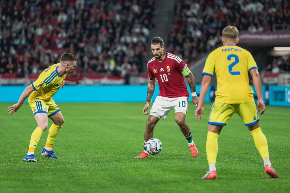 Trei jucători de fotbal sunt pe un teren de iarbă verde, doi purtând echipamente galbene și unul purtând o echipă roșie cu pantaloni albi.
