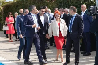 Un grup de oameni în haine formale merge pe o suprafață pavată lângă un bazin, în timp ce un bărbat filmează cu o cameră video.