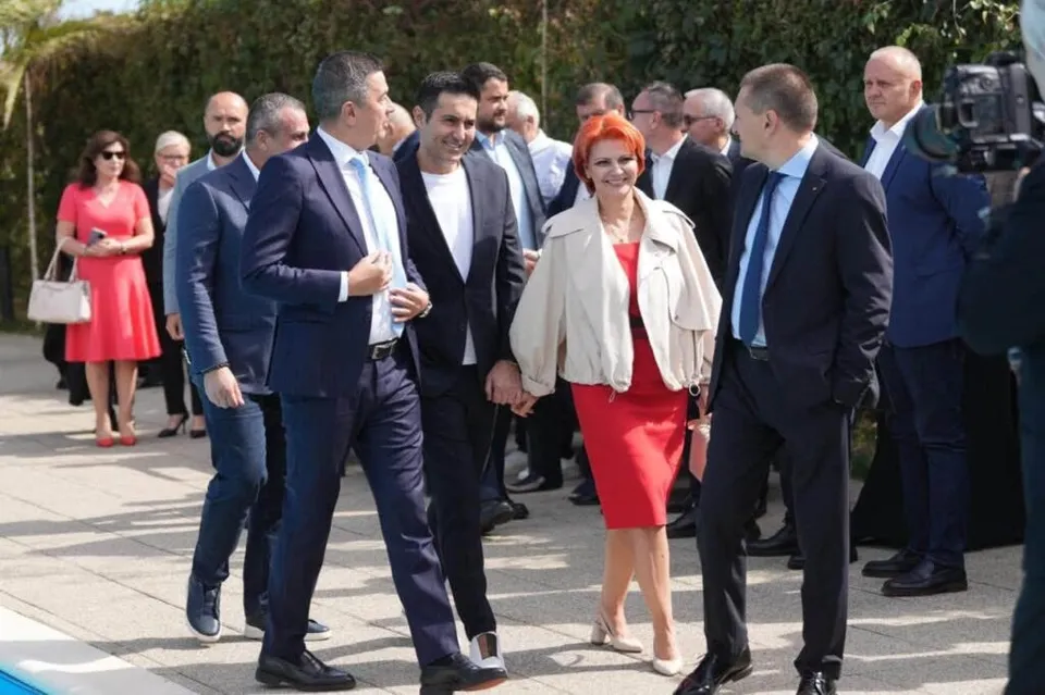 Un grup de oameni în haine formale merge pe o suprafață pavată lângă un bazin, în timp ce un bărbat filmează cu o cameră video.