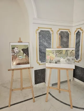 Două ilustrații sunt expuse pe easete de lemn într-o încăpere cu pereți albi și panouri de marmură în fundal.