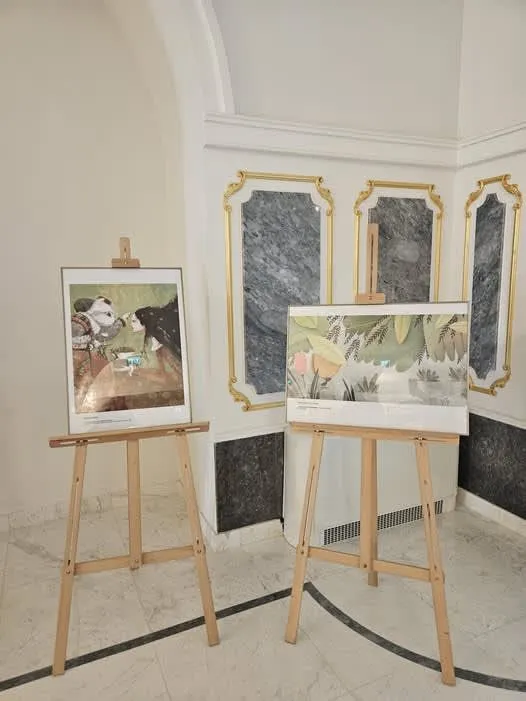 Două ilustrații sunt expuse pe easete de lemn într-o încăpere cu pereți albi și panouri de marmură în fundal.
