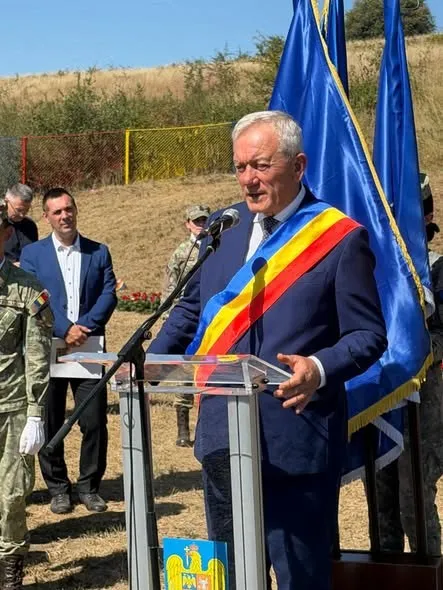Un bărbat într-un costum albastru cu o bandă tricoloră pe umăr vorbește la un microfon aflat pe un podium transparent în fața unor steaguri albastre.