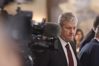 Un bărbat cu părul gri, purtând costum închis și cravată roșie, este filmat de o cameră video în prim-plan.