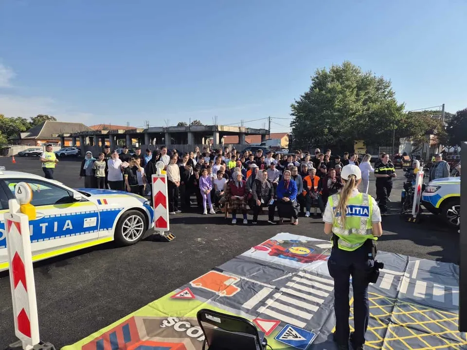 Un grup de persoane stă adunat pe un teren asfaltat lângă mașini de poliție și un covor colorat cu marcaje rutiere.