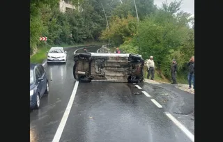 Un autoturism alb este răsturnat pe o laterală pe un drum asfaltat, în timp ce alți șoferi și persoane stau pe marginea drumului.