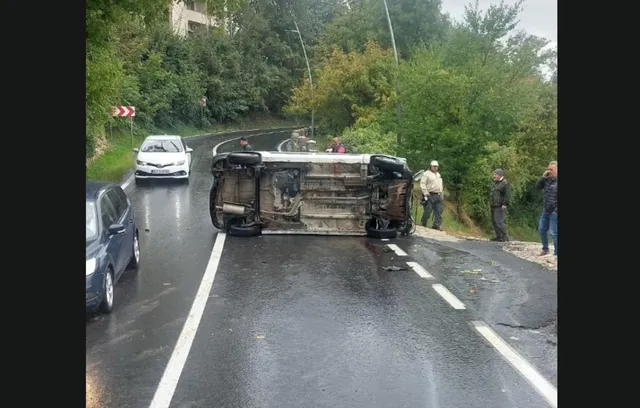 Un autoturism alb este răsturnat pe o laterală pe un drum asfaltat, în timp ce alți șoferi și persoane stau pe marginea drumului.