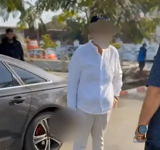 Un bărbat cu cămașă albă și pantaloni deschiși stă lângă o mașină gri pe un drum asfaltat.