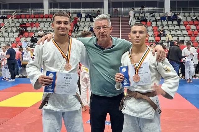 Trei bărbați stau în picioare pe un covor de sport, doi purtând kimono albe și medalii cu pangalii colorate, ținând diplome de carton.