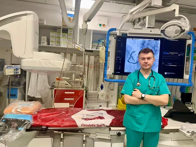 Un bărbat în halat medical verde stă cu brațele încrucișate în fața unui monitor medical într-o încăpere de spital dotată cu echipamente tehnice.