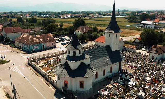 O vedere aeriană prezintă o biserică albă cu acoperiș închis și un cimitir cu morminte situate lângă o stradă și case.