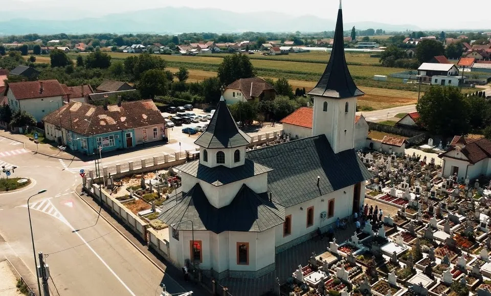 O vedere aeriană prezintă o biserică albă cu acoperiș închis și un cimitir cu morminte situate lângă o stradă și case.