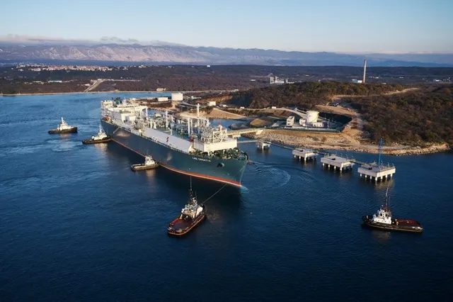 O navă mare de transport de gaz este înconjurată de patru remorchere în apă, lângă o instalație industrială de pe malul unui corp de apă.
