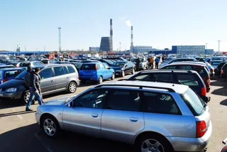 Mulți autoturisme sunt parcate pe o suprafață mare de asfalt sub un cer senin, având turnuri industriale în fundal.