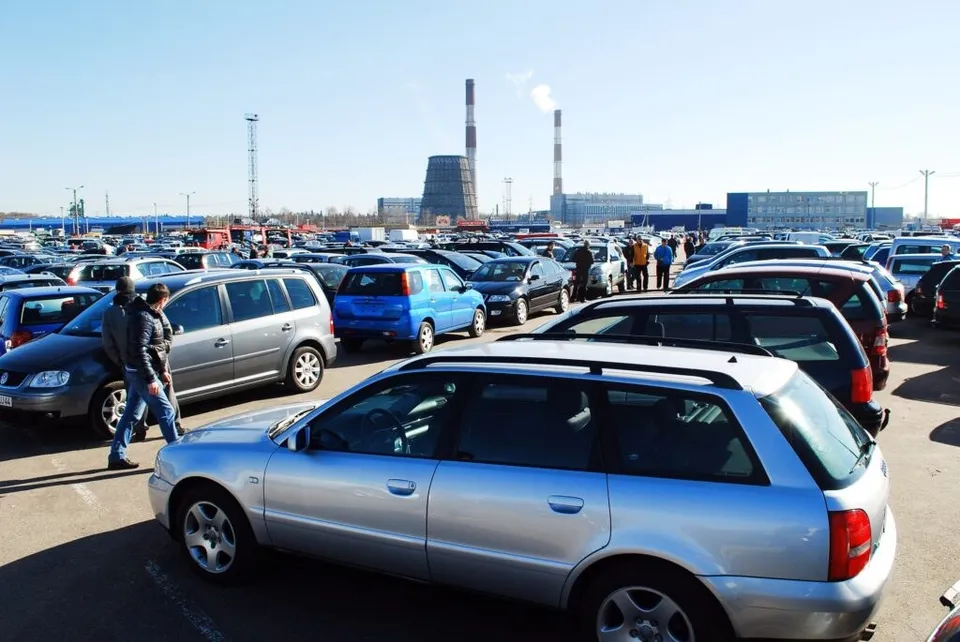Mulți autoturisme sunt parcate pe o suprafață mare de asfalt sub un cer senin, având turnuri industriale în fundal.