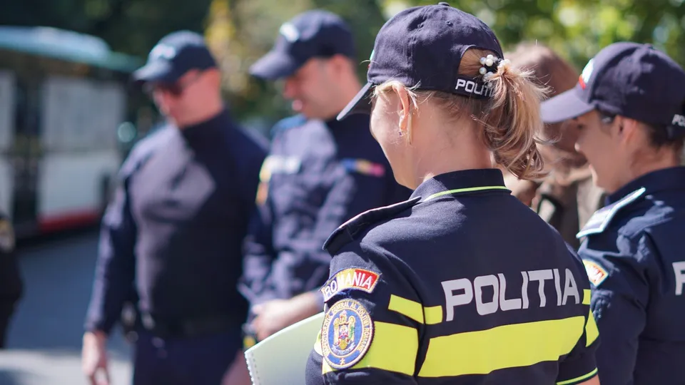 Trei polițiști, doi bărbați și o femeie, sunt fotografiați din spate, în uniformă, pe stradă, cu un autobuz în fundal.
