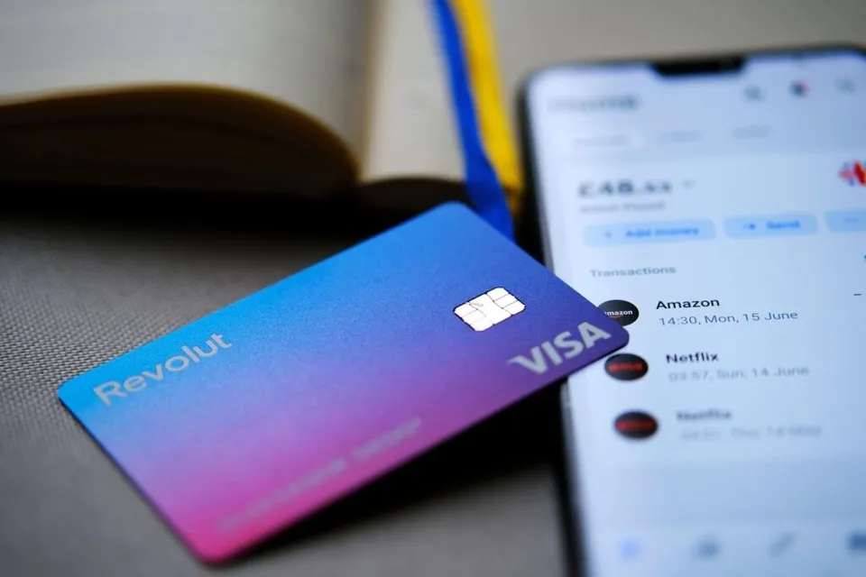 O carte de credit albastră și mov cu logo-ul Revolut și Visa este așezată lângă un telefon mobil care afișează o listă de tranzacții.