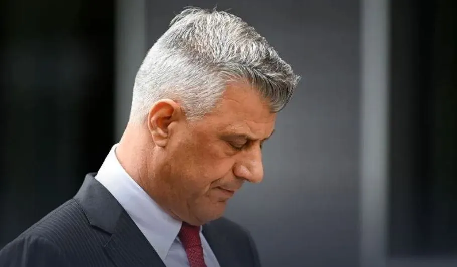Un bărbat cu păr cărunt, îmbrăcat la costum cu cravată roșie, privește în jos, cu o expresie serioasă.