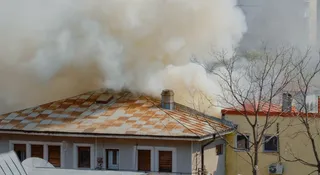 Fum dens și alb se ridică deasupra acoperișului unei clădiri cu țiglă ruginită lângă un copac fără frunze.