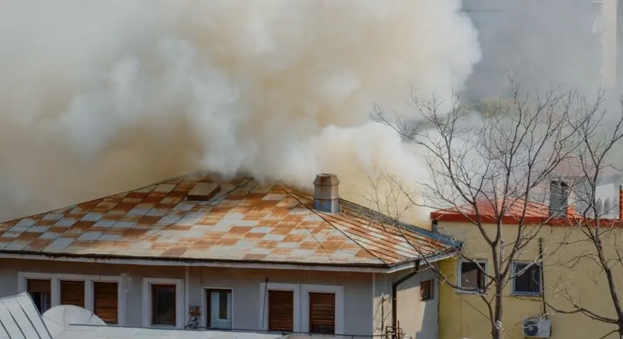 Fum dens și alb se ridică deasupra acoperișului unei clădiri cu țiglă ruginită lângă un copac fără frunze.