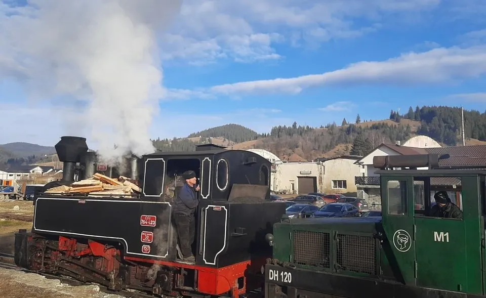 Un tren cu abur de culoare neagră și roșie trage vagoane pe o cale ferată, emanând fum alb sub un cer parțial înzăpezit.