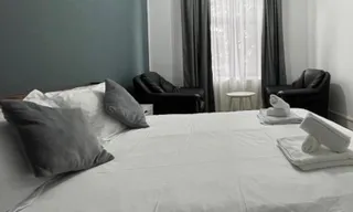 Un pat cu lenjerie albă, două perne gri și două seturi de prosoape albe alese sunt așezate pe suprafața acestuia într-o cameră cu fotolii negre.