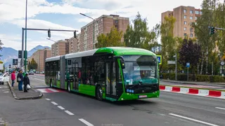 Un autobuz articulat de culoare verde circulă pe un drum asfaltat lângă un trotuar unde stau câțiva oameni.
