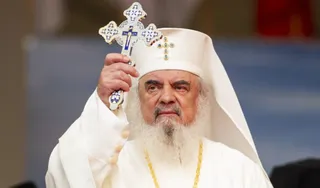 Un bărbat cu barbă albă și pălărie albă ține un cruce metalică decorată cu pietre albastre.