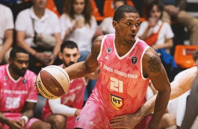 Un jucător de baschet cu mânecă de echipă roz și numărul 21 conduce o minge de baschet pe un teren de sport.