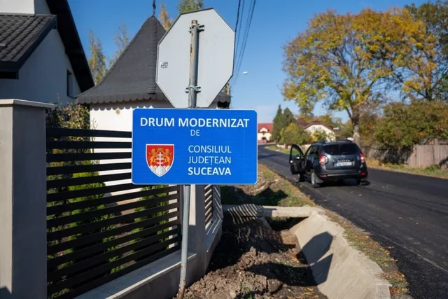 O placă albastră cu textul „DRUM MODERNIZAT DE CONSILIUL JUDEȚEAN SUCEAVA” este montată pe un stâlp lângă un drum asfaltat și o mașină parked.