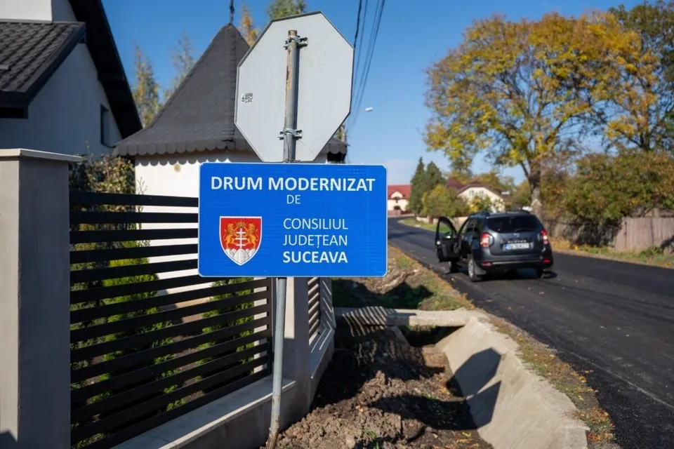 O placă albastră cu textul „DRUM MODERNIZAT DE CONSILIUL JUDEȚEAN SUCEAVA” este montată pe un stâlp lângă un drum asfaltat și o mașină parked.