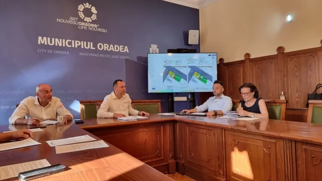 Patru persoane stau la o masă de conferințe din lemn sub un ecran care afișează două hărți colorate.