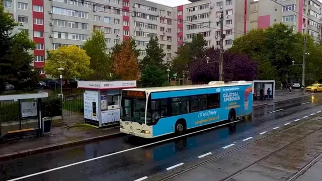 Un autobuz albastru și alb circulă pe un drum umed lângă un autobuz și un shelter de așteptare, având blocuri de apartamente în fundal.