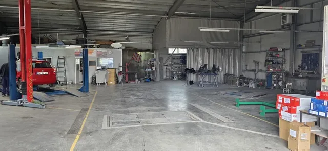 Interiorul unui atelier auto cu un lift roșu, un autoturism roșu ridicat și diverse unelte și cutii pe podea.