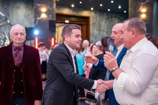 Un bărbat într-un costum negru strânge mâna unui bărbat într-o cămașă albă, în timp ce alți oameni stau în grup în fundal.