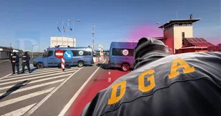 O persoană cu o vestă neagră pe care scrie DGA în litere galbene stă în prim-plan, în fața unor mașini de poliție și a unui punct de frontieră.