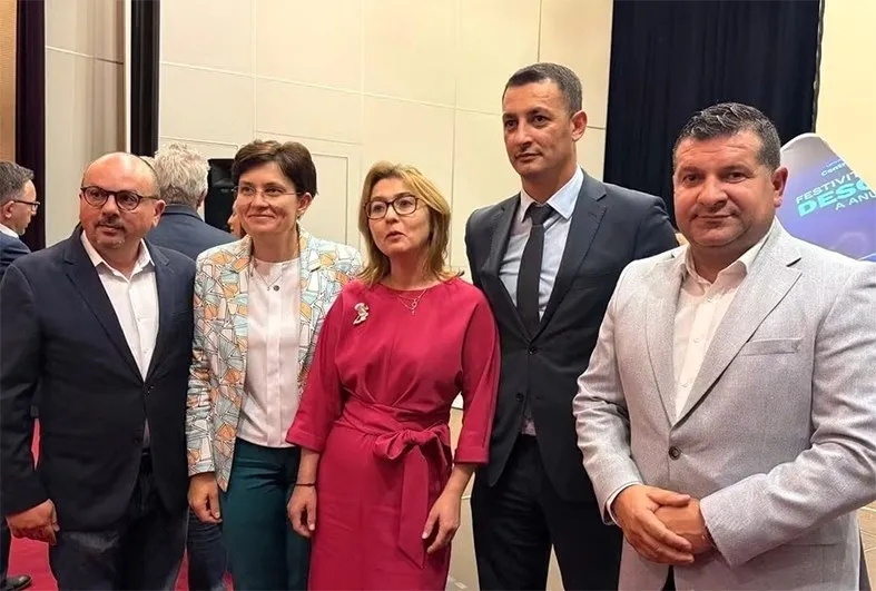 Cinci persoane stau în picioare într-o încăpere interioară, purtând haine formale, în fața unui fundal cu panouri deschise și un panou albastru.